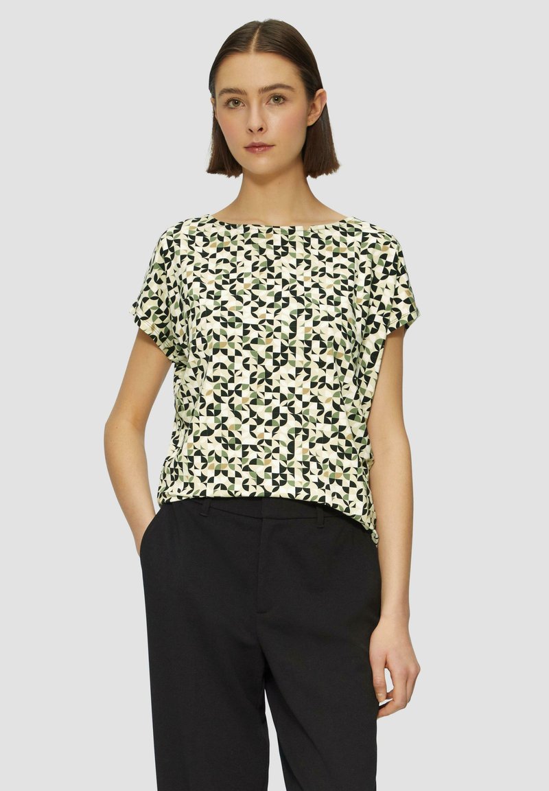 Korte mouw top in crèmekleurige stof met een geometrisch patroon in zwart, groen en bruin. Eenvoudige ronde hals en een relaxte pasvorm. Gepaard met zwarte broek.