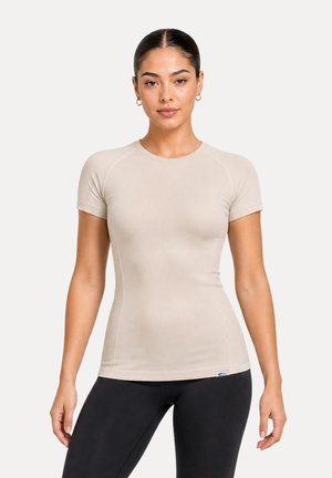 Femme aux cheveux foncés portant un haut beige ajusté à manches courtes et des leggings noirs, debout face à un fond clair uni.
