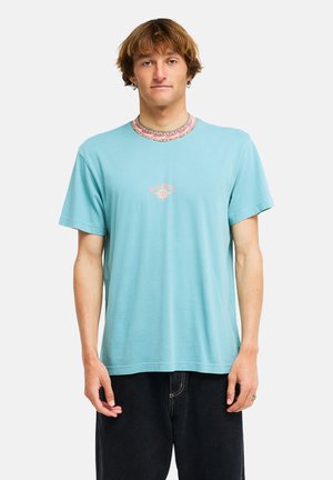 Jonge man met kort bruin haar, met een lichtblauw T-shirt met kleine print op de borst en zwarte broek, staand tegen een witte achtergrond.