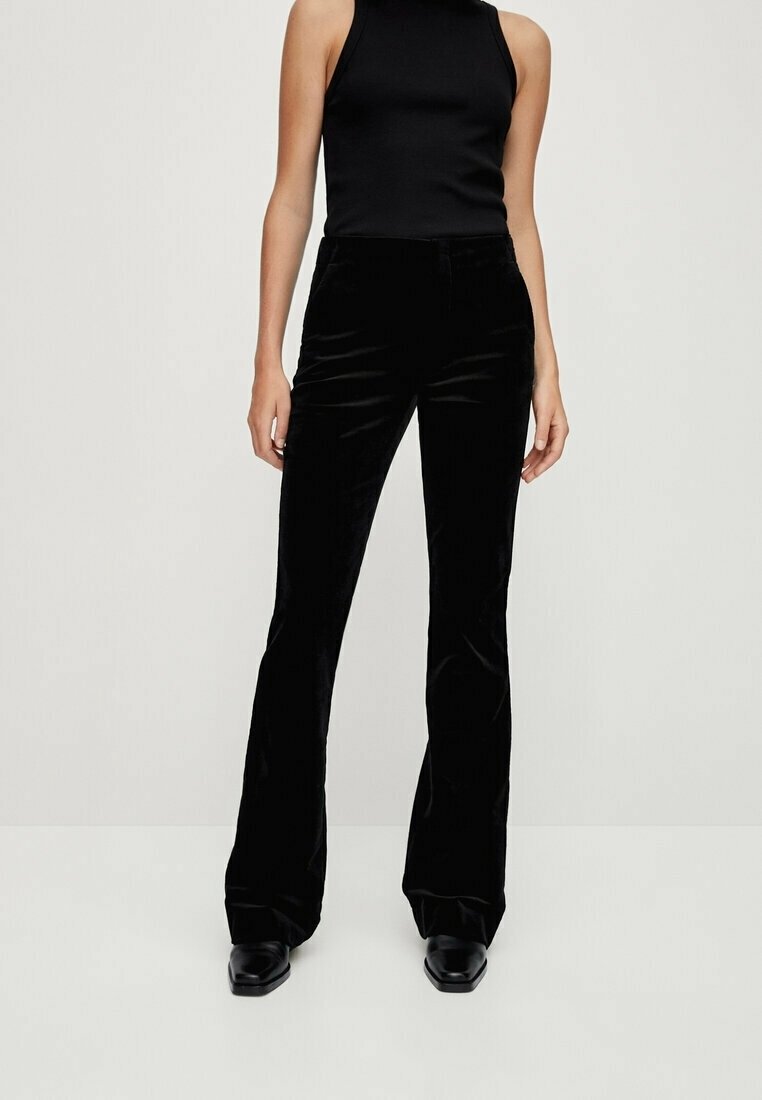 Femme portant un haut noir sans manches, un pantalon évasé en velours noir et des chaussures noires, debout devant un fond clair uni.
