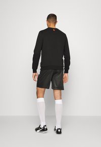 Felpa nera con logo rosso, indossata con pantaloni sportivi neri e calze a ginocchio bianche, caratterizzata da un tessuto testurizzato e una vestibilità comoda.