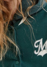 Sudadera verde de textura suave, con un logotipo blanco en forma de "M" y cierre de botones. Incluye una capucha de doble capa con detalles a rayas.