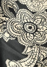 Črn material z cvetličnim vzorcem v krem barvi, z zapletenimi spiralami in konturami. Oblikovanje vključuje drzne, jasno definirane oblike in akcentne dele.