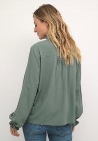 Blusa verde de manga larga en tejido ligero con un corte relajado, detalle fruncido en los puños y un yugo en la parte posterior. Con jeans azules.