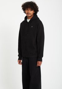 Black zip-up hoodie made of soft fabric, featuring a front pocket and adjustable drawstrings. Paired with wide-legged black pants.