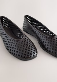 Chaussures plates noires slip-on avec perforations carrées sur toute la surface, sur un fond clair uni.