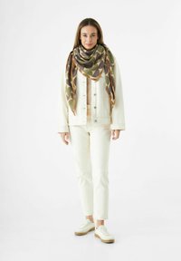 Femme debout portant un jean blanc, une veste crème, une écharpe oversize à motifs verts et bruns, et des baskets blanches sur fond blanc.