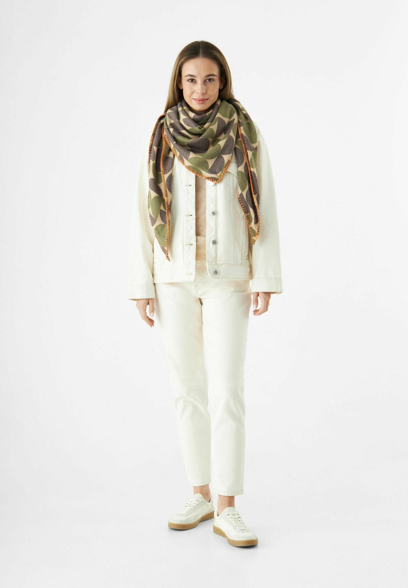 Femme debout portant un jean blanc, une veste crème, une écharpe oversize à motifs verts et bruns, et des baskets blanches sur fond blanc.