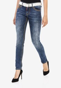 Schlanke blaue Jeans aus Denim mit heller Verwaschung, kontrastierenden Nähten und einem weißen, mit Nieten besetzten Gürtel. Das Model trägt schwarze High Heels.