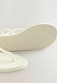Weiße Flip-Flops mit einer strukturierten Gummisohle und dünnen, flexiblen Riemen, die kleine Perlen entlang des Zehenbalkens und eine glatte Fußbett haben.