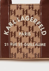 Hnědá plátěná taška s opakujícím se vzorem loga v tmavě hnědé a béžové barvě. Bílé písmo zobrazuje "KARL LAGERFELD," "PAŘÍŽ," a adresu.