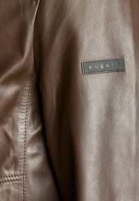 Bruine leren jas met een subtiele textuur. Inclusief een zwart label met "bugatti" erin geperst, gestikte details langs de naad.