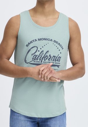 Mann trägt ein hellgrünes ärmelloses Shirt mit dem Text „Santa Monica Beach California“ und blaue Jeans, Hände vor dem Körper verschränkt.