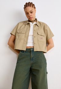 Cămașă cropped bej, cu mâneci scurte, buzunare pe piept și închidere cu nasturi, asortată cu pantaloni din denim verzi, cu talie înaltă și croi lejer.