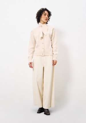 Light pink knitted cardigan with large buttons and a bow tie collar, paired with wide cream trousers and dark ankle boots.