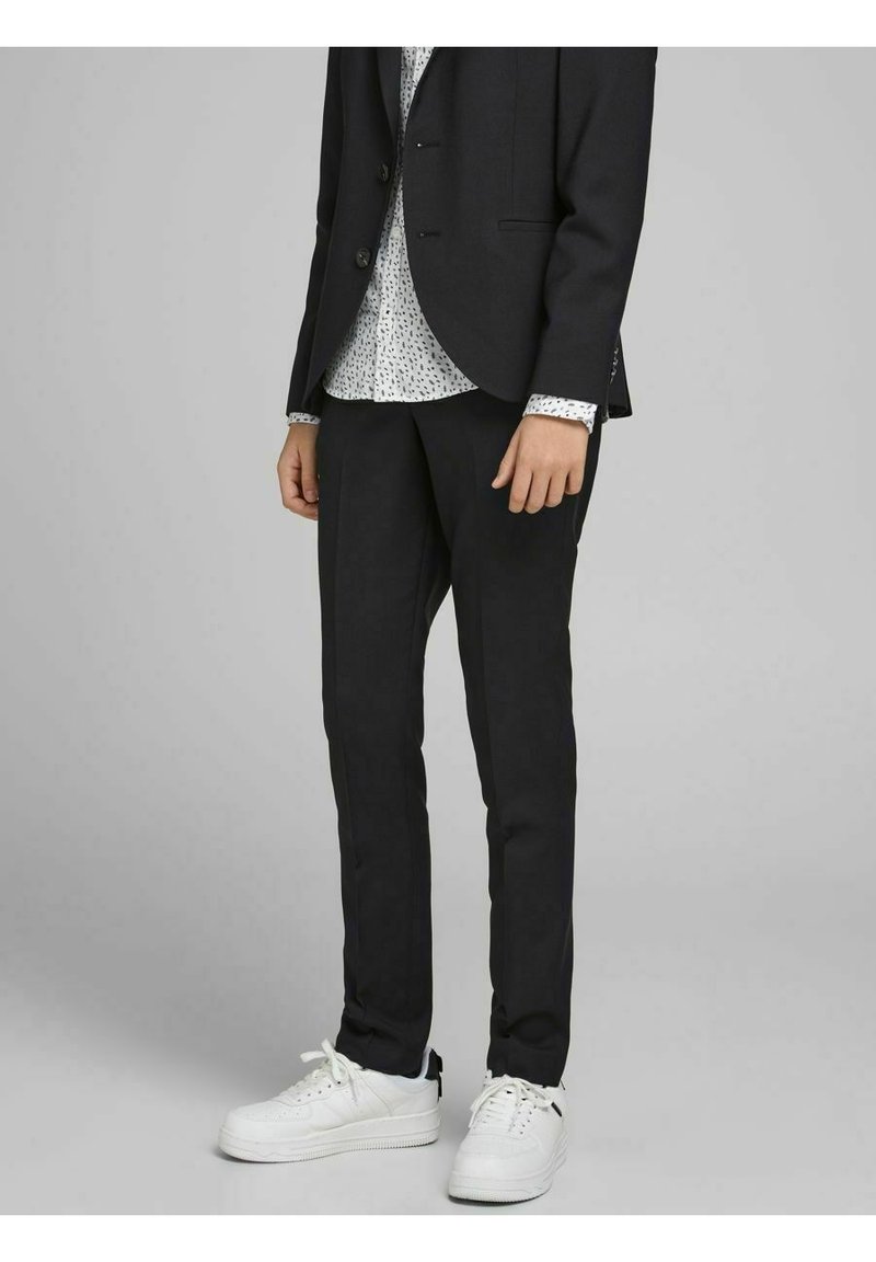 Personne portant un pantalon de costume noir, un blazer noir, une chemise blanche à motifs et des baskets blanches, debout devant un fond gris.