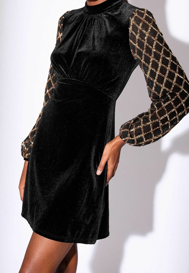 Robe courte en velours noir avec taille froncée, col montant et manches longues transparentes avec motif en treillis doré. Pose avec la main sur la hanche.