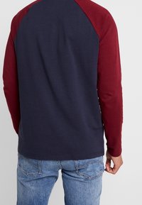 Pitkähihaisen t-paidan navy-värinen vartalo ja viininpunaiset raglan-hihat, valmistettu pehmeästä kankaasta, jonka pinta on sileä. Rentoa suunnittelua suoran helman kanssa.