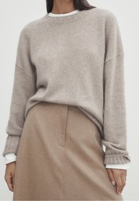 Pull en tricot beige avec un col rond et des poignets côtelés, associé à une jupe en laine beige présentant une poche latérale et une silhouette épurée.