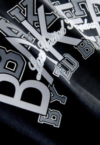 Sort t-shirt i stof med lagdelt hvid og sølv tekst i forskellige skrifttyper, der viser ordene "BAKER" og "London United Kingdom".