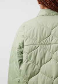 Jachetă verde matlasată cu o textură netedă și un model de valuri. Prezentă un guler și o croială lejeră, evidențiind zonele umplute.