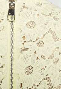 Krämfärgad blommig spets med en silverdragkedja märkta "BOSS" som löper vertikalt på vänster sida.