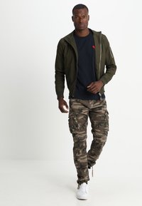 Olijf groene ritsjas over een marineblauw T-shirt, gecombineerd met camouflage cargo-broeken en witte sneakers, met meerdere zakken en een comfortabele pasvorm.