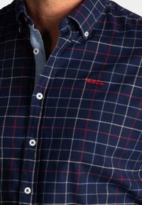 Marineblauw geruit shirt met rode en witte vierkanten, met knoopsluiting en geborduurd logo in het rood. Heeft witte knopen en een zachte textuur.