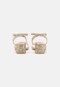Glittriga guld högklackade sandaler med en fyrkantig blockklack, korsade remmar och en slät beige sula.