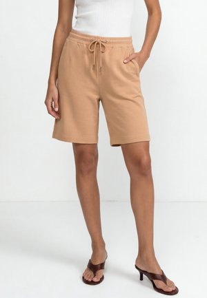 Vrouw draagt beige short met trekkoord, wit mouwloos topje en bruine sandalen met hak, staand tegen een effen witte achtergrond.
