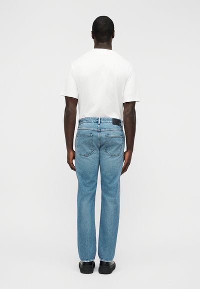 Ανοιχτό μπλε τζιν με ίσια γραμμή, με δερμάτινο ταμπελάκι στη μέση πίσω και δύο πίσω τσέπες. Συνδυασμένο με λευκό T-shirt.