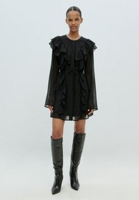 Schwarzes Kleid mit Rüschendetails, langen durchsichtigen Ärmeln und tailliertem Bund. Kombiniert mit knielangen, glänzenden schwarzen Stiefeln.