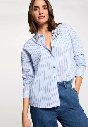 Mujer joven con una camisa de botones a rayas azules y blancas con cuello adornado y jeans azules, posando con una mano en el bolsillo.