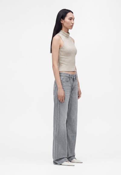 Jeune femme debout de profil portant un top sans manches à col roulé rayé, un jean gris clair à jambes larges et des chaussures blanches à bouts pointus.