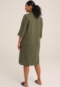 Abito verde oliva con una vestibilità rilassata, maniche a tre quarti e tessuto testurizzato. L'orlo arriva sotto il ginocchio con spacchi laterali.