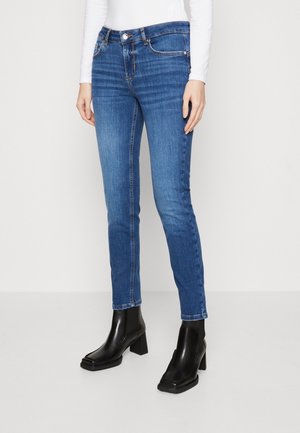 Kvinde iført tætsiddende blå denimjeans, hvid langærmet top og sorte støvler med hæl, står mod en ensfarvet lys baggrund.