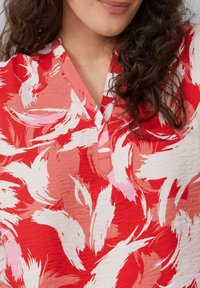 Rode en witte geblokte blouse met een V-hals, textuurstof en een abstracte penseelstreekontwerp met variërende tinten roze.