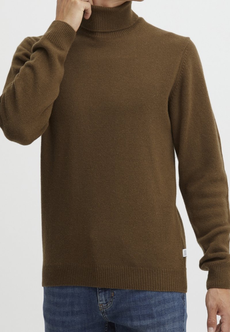 Pull en maille brune à col roulé avec poignets et ourlet côtelés. Texture douce, coupe légèrement ajustée, associé à un jean denim bleu.