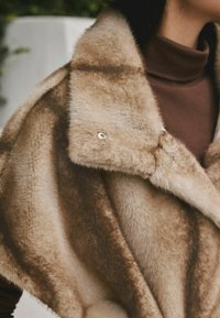 Faux fur shawl in shades of beige with a soft texture. Features a large collar and a silver button accent for closure.