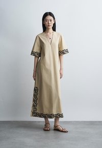 Femme debout dans une robe midi beige avec une broderie circulaire noire sur les manches, l'ourlet et le côté, portant des sandales marron et un collier avec pendentif.