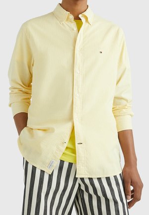 Homme portant une chemise jaune clair à fines rayures boutonnée par-dessus un t-shirt jaune et un pantalon à rayures verticales noir et blanc.