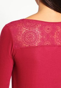 Burgundy topp med en texturerad spetsbakgrund med cirkulära mönster och rund halsringning. Tyget är slätt och har en figurnära silhuett.