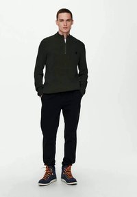 Pull zippé vert foncé à texture côtelée, associé à un pantalon noir et des baskets montantes bleues avec des lacets orange. Design simple, coupe décontractée.