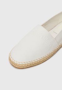 Bijela platnena espadrila s prirodnim jutom, s reljefnim detaljem logotipa na gornjem dijelu i šivanim rubom oko potplata.