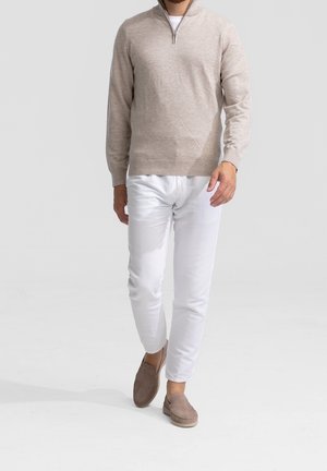 Homme marchant portant un pull beige à demi-fermeture éclair, pantalon blanc et chaussures beige à enfiler sur un fond clair uni.