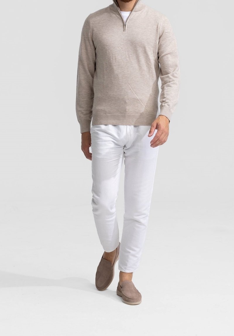 Homme marchant portant un pull beige à demi-fermeture éclair, pantalon blanc et chaussures beige à enfiler sur un fond clair uni.