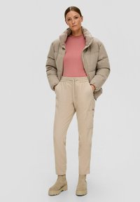 Beige pufferjack met hoge kraag over een roze geribbelde top, gecombineerd met crèmekleurige cargobroek en beige enkellaarsjes. Zachte texturen, relaxte pasvorm.