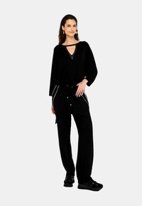 Top nero con scollatura a V e maniche lunghe, abbinato a pantaloni neri con tasche laterali e accenti bianchi, indossato con sneakers nere.