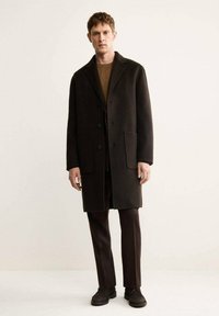 Cappotto di lana scuro con colletto a punta, tre bottoni e due tasche frontali. Indossato sopra un maglione marrone e abbinato a pantaloni scuri e calzature.