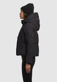 Sort, quilted pufferjakke med høj krave og hætte. Har en sidelomme og en rund logoapplikation på ærmet.